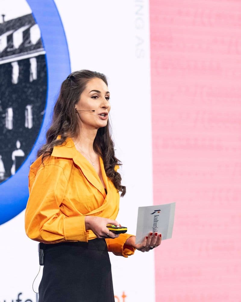 Hana Colakovic på scenen i oransje bluse, med Kulturjakten-kort i hånden